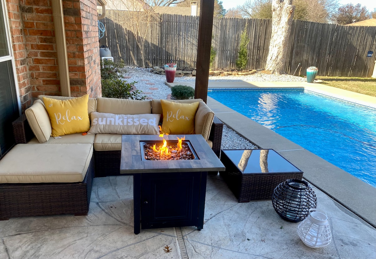 An outdoor seating area features a cushioned sectional sofa arranged around a fire pit, creating a cozy gathering space. A clear blue pool is visible in the background, complemented by a well-maintained yard and a wooden privacy fence.