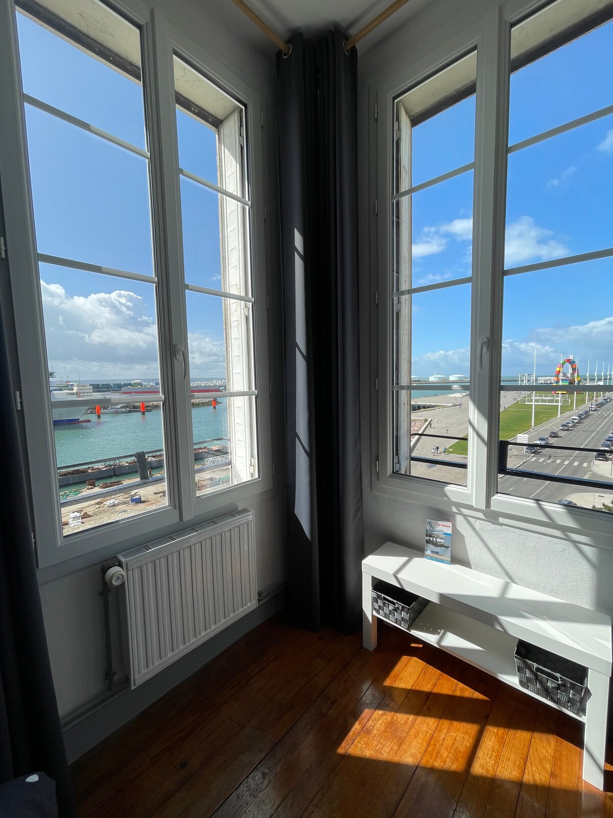 Large windows frame a stunning view of the harbor and vibrant waterfront, allowing abundant natural light to fill the space. The polished wooden floor and minimalistic white furniture create a clean and welcoming atmosphere, emphasizing the connection to the outdoor scenery.
