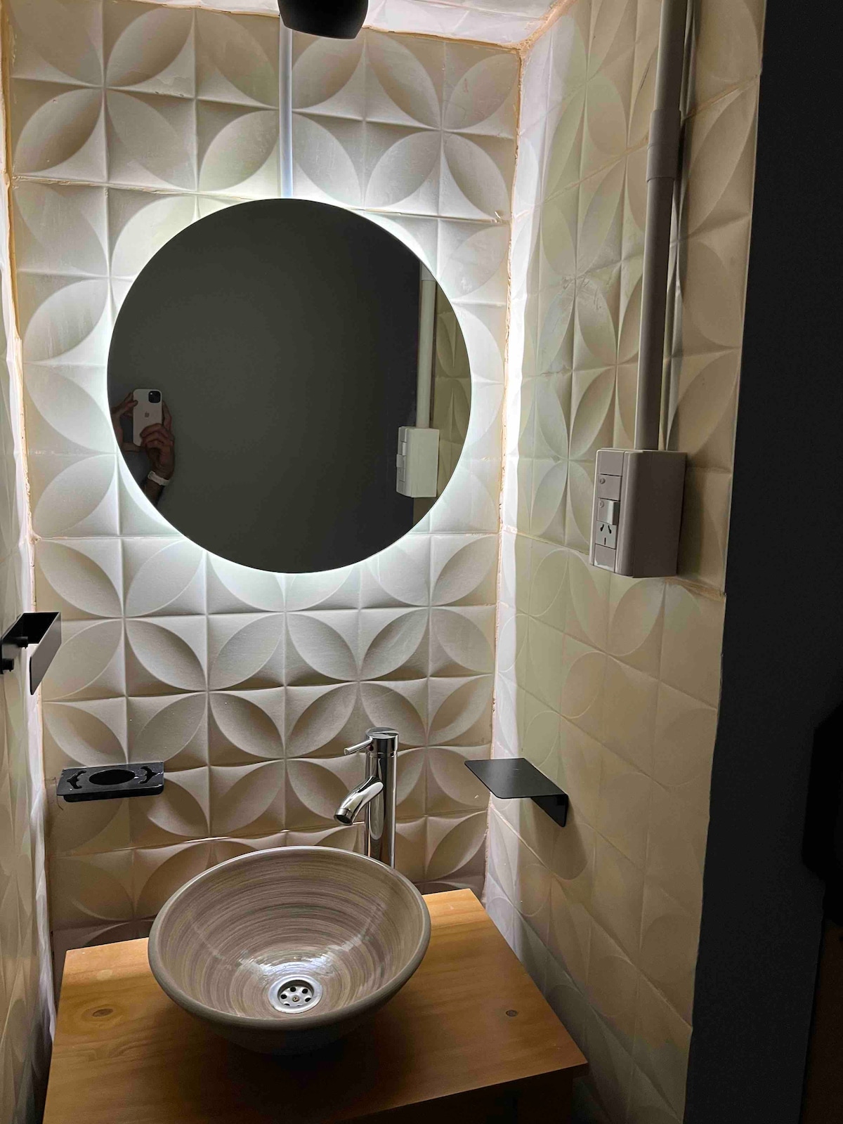 A modern bathroom corner is shown, featuring a round mirror that is softly illuminated. A circular, textured sink rests on a wooden surface, complemented by minimalist shelving. Intricate patterned tiles line the walls, enhancing the space's contemporary design.