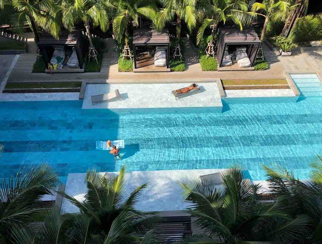 An aerial view captures a serene swimming pool surrounded by tropical greenery. Lounge chairs are positioned alongside the pool, while shaded cabanas provide relaxation areas. The water's surface reflects the sunlight, enhancing the inviting atmosphere of the outdoor space.