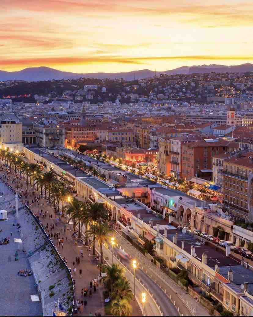 An elevated view of a vibrant coastal promenade at sunset, showcasing lined palm trees and bustling streets filled with people. The horizon is painted in warm tones, with hills viewed in the background, emphasizing the lively atmosphere of the waterfront.