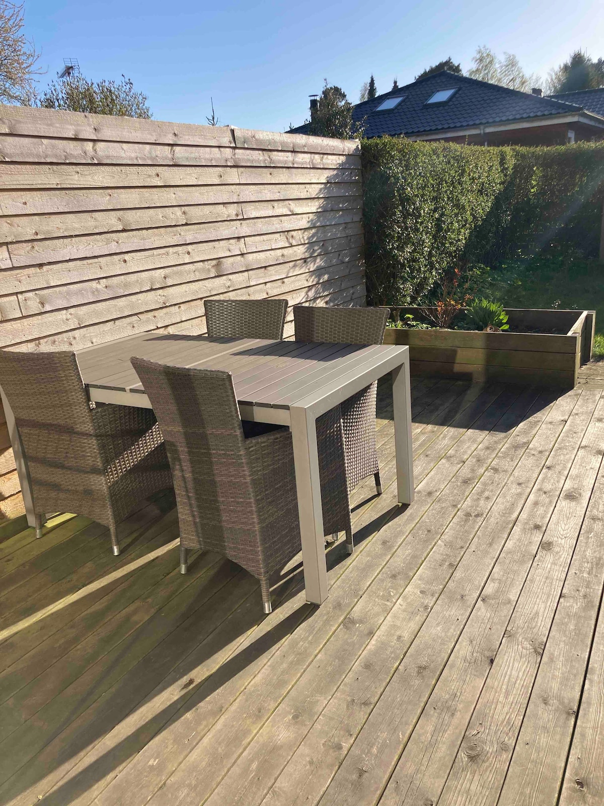 An outdoor terrace features a wooden deck with a dining table and four woven chairs. A raised planter and hedge line the area, providing a sense of privacy. Natural light enhances the inviting atmosphere of this outdoor space.