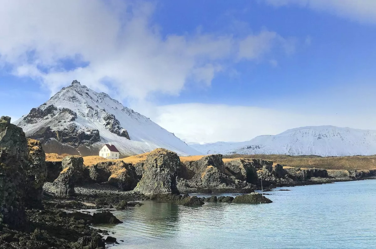 A serene landscape showcases rugged coastal rocks meeting calm waters. In the distance, a cottage is positioned against a backdrop of snow-covered mountains and a bright blue sky, highlighting the natural beauty of the Snæfellsnes peninsula.