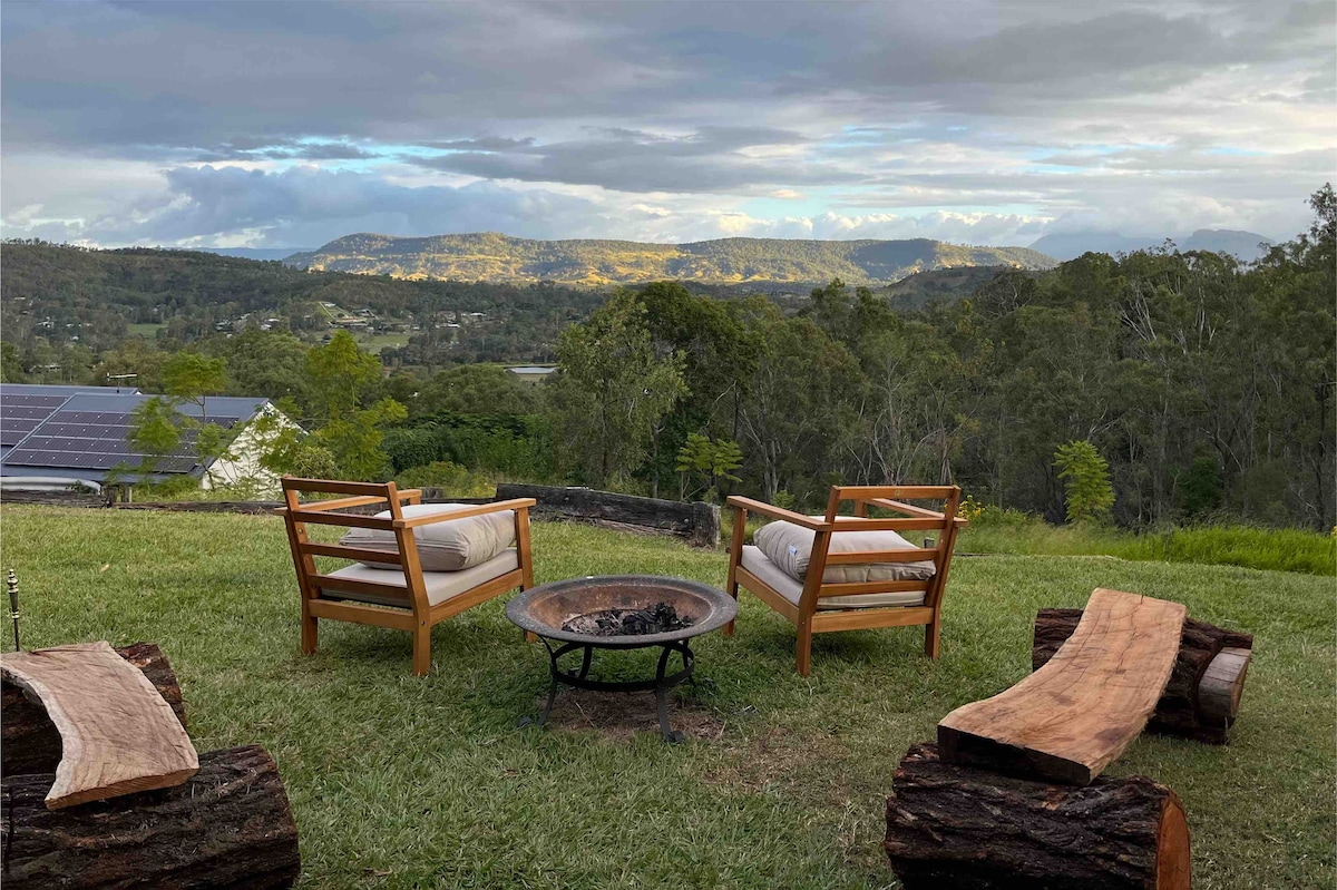 Two wooden chairs are positioned around a fire pit, surrounded by log seating. The view captures the expansive Great Dividing Range in the distance, framed by lush greenery and an open lawn area under a partly cloudy sky.