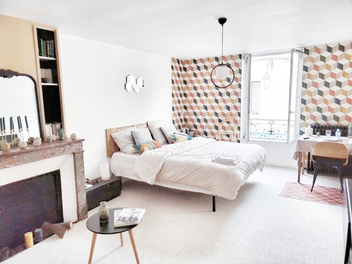 A spacious sleeping area features a double bed with soft bedding. Geometric patterned walls provide a modern touch. Natural light enters through a window, while a small round table complements the space. A decorative mirror and a cozy rug enhance the inviting ambiance.
