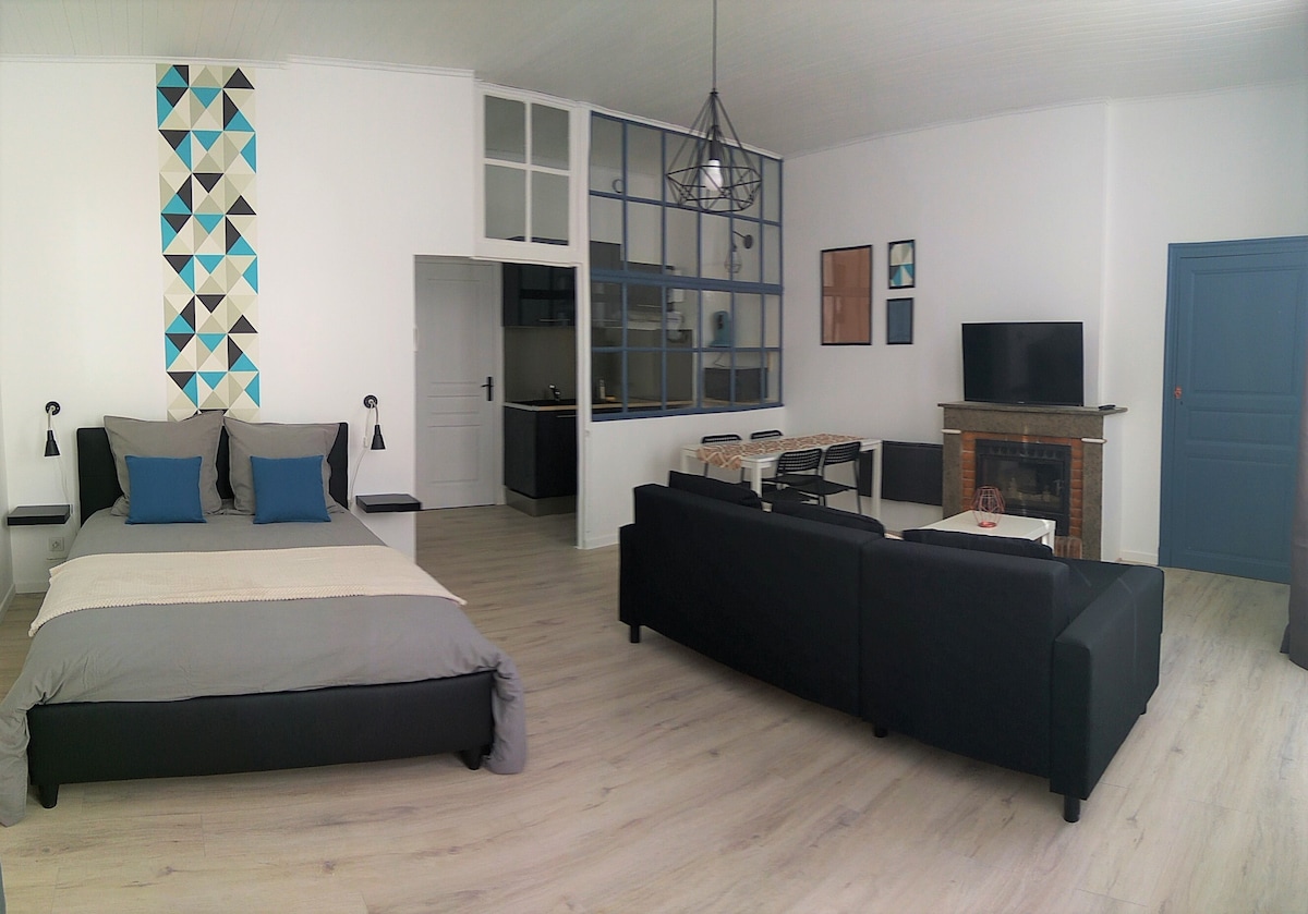 A spacious studio features a comfortable bed with two pillows and a textured blanket. A modern kitchen area is visible in the background, while a flat-screen television is mounted on a wall opposite a cozy seating arrangement. Decorative wall art adds a pop of color.