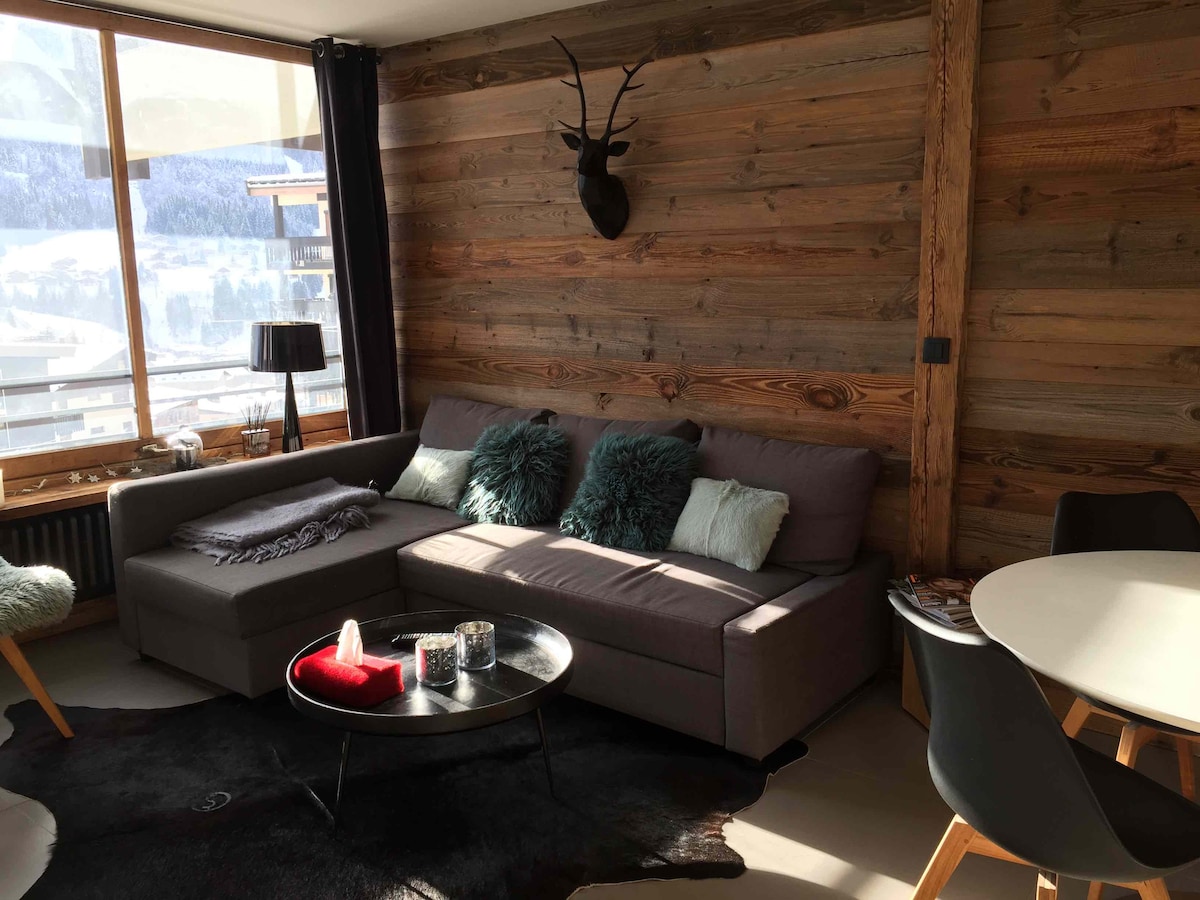 The living area features a large sectional sofa with plush cushions, positioned near a window that reveals snowy mountain views. A circular coffee table stands in front of the sofa, with decorative items arranged on it. A dining table with modern chairs occupies the corner, enhancing the open space.