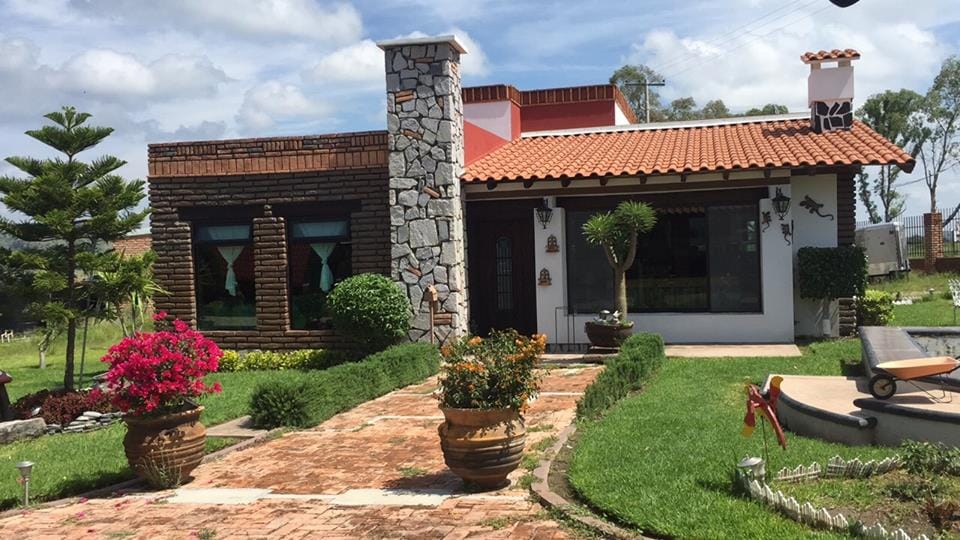 A charming cabin exterior is viewed featuring a stone chimney and a rustic tile roof. Seasonal flower pots are positioned along a pathway, surrounded by lush green grass and shrubbery. Large windows are framed by decorative curtains, enhancing the inviting appearance of the structure.