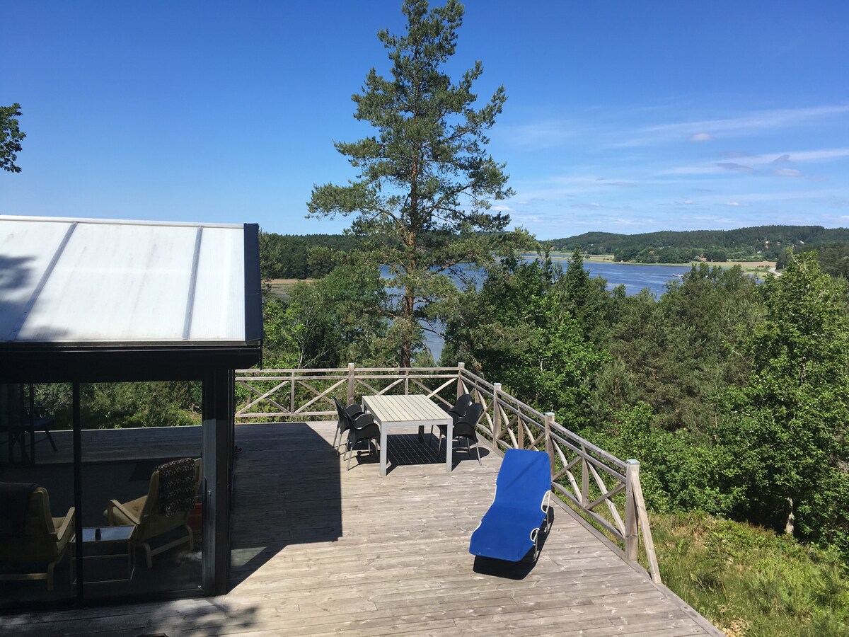A spacious wooden deck features lounge chairs and a dining table, surrounded by lush greenery. The view opens up to a serene landscape of water and distant hills under a clear blue sky, creating a peaceful setting for relaxation.