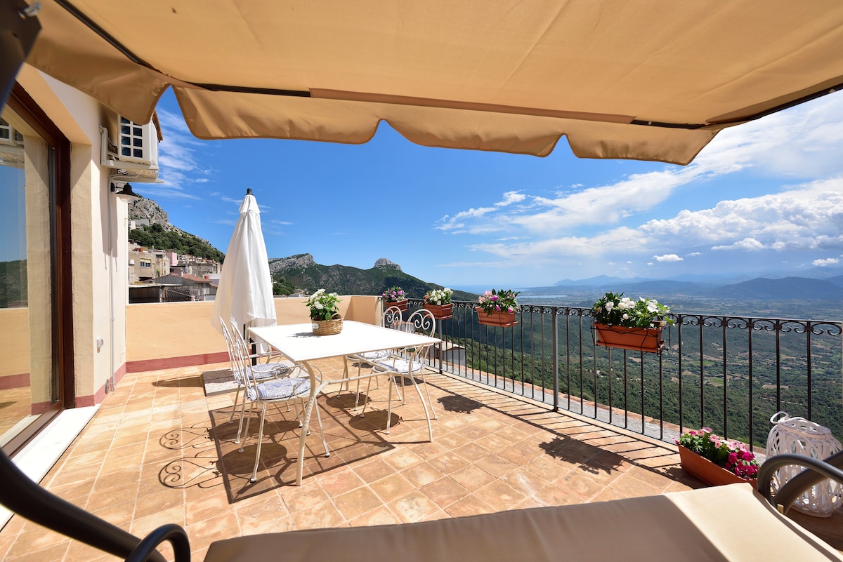A spacious terrace is featured, furnished with a dining table and surrounded by flowering plants in pots. An awning provides shade, while expansive views of the mountains and valley are visible in the distance under a clear blue sky.