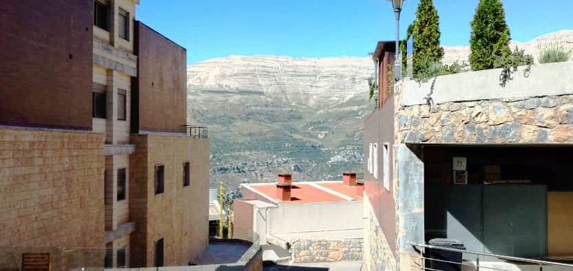 A scenic view of Zaarour Hills is captured, featuring terraced buildings that show a blend of stone and modern architecture. The mountainous landscape stretches beyond, under a clear blue sky, providing a sense of tranquility and connection to nature.