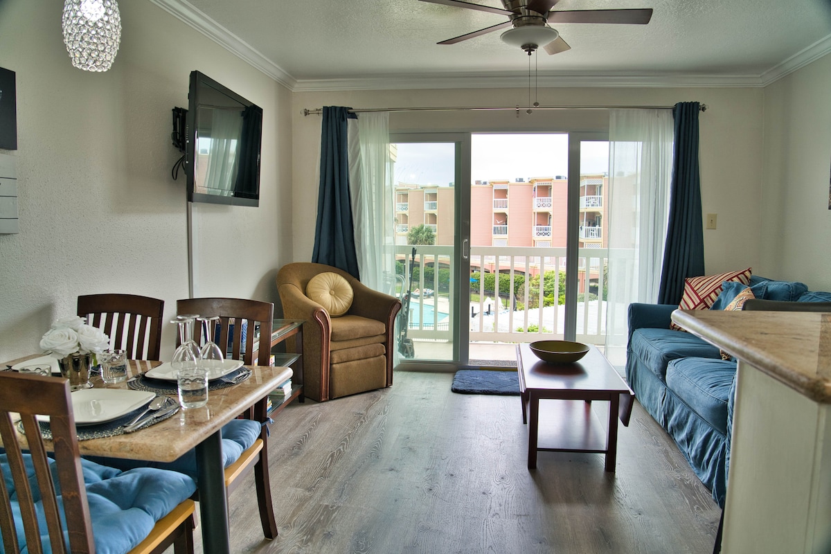 A living area features a comfortable blue couch and a beige armchair, positioned near large sliding glass doors that open to a patio with a view of the pool. A dining table with four chairs sits adjacent, complemented by a flat-screen TV mounted on the wall.