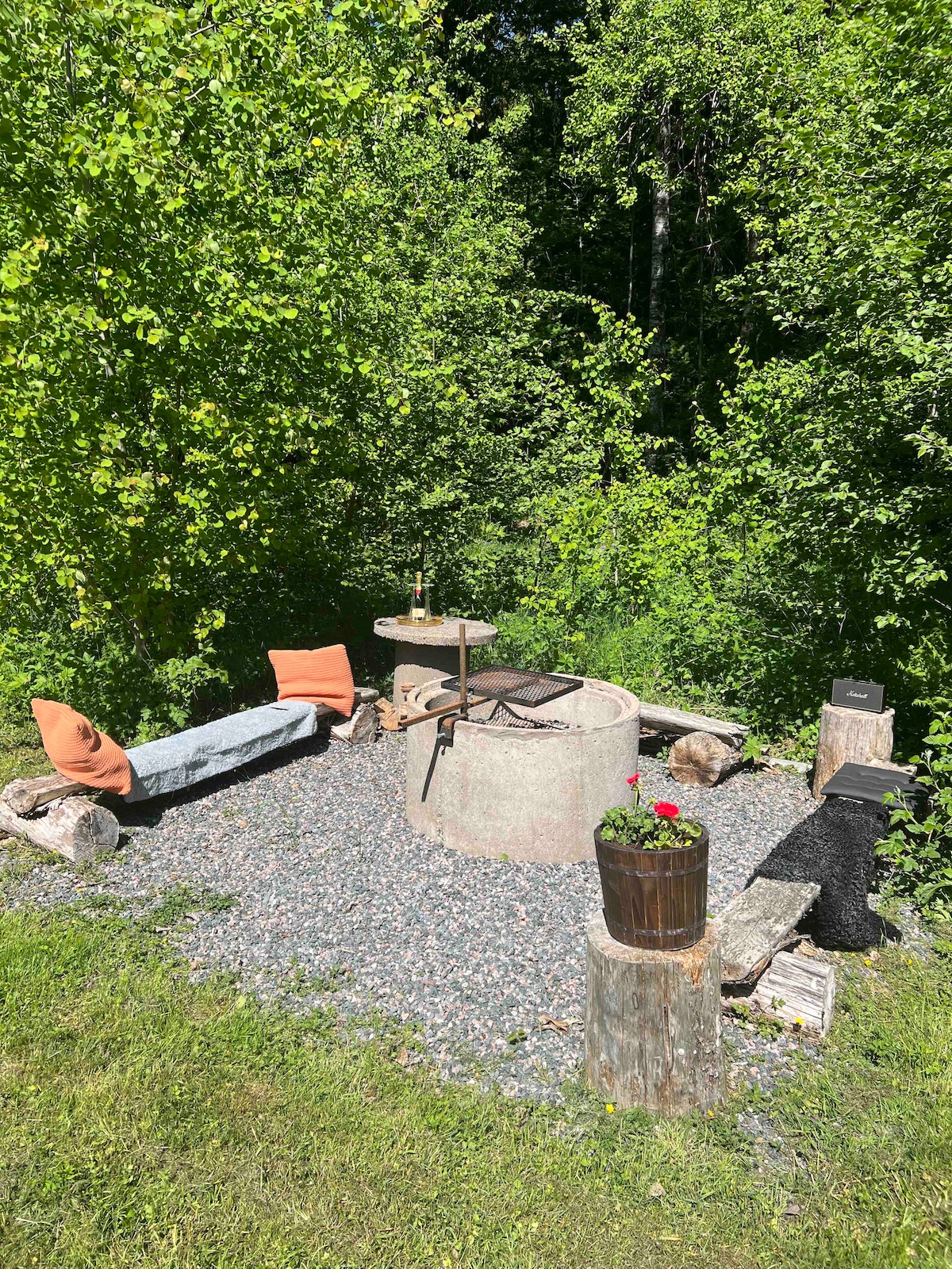 A cozy outdoor seating area is featured, surrounded by lush greenery. A circular fire pit sits at the center, with logs and stones creating a rustic seating arrangement. Colorful pillows add comfort, while a flower pot brings a touch of color to the natural setting.