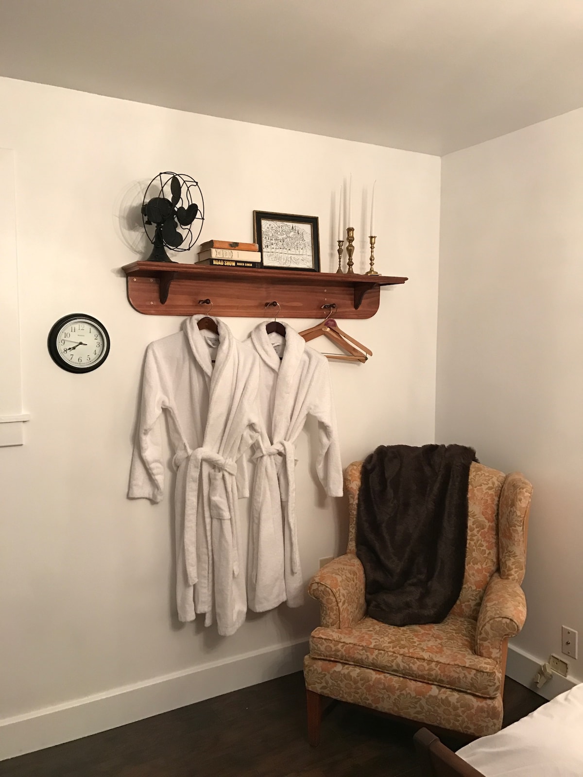 A white shelf is mounted on the wall, displaying an old-fashioned fan, decorative candles in holders, and a framed artwork. Two plush white robes hang below it on hooks. An ornate upholstered chair accompanied by a soft throw is positioned nearby, with a round clock visible on the wall.