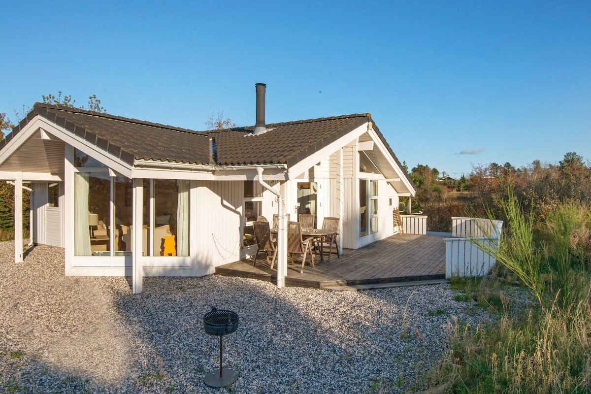 The cottage features a modern exterior with a sloped roof and large windows, allowing for ample natural light. A spacious wooden deck is visible, furnished with outdoor seating and a grill. Surrounding greenery complements the serene setting, with a gravel area leading to the entrance.