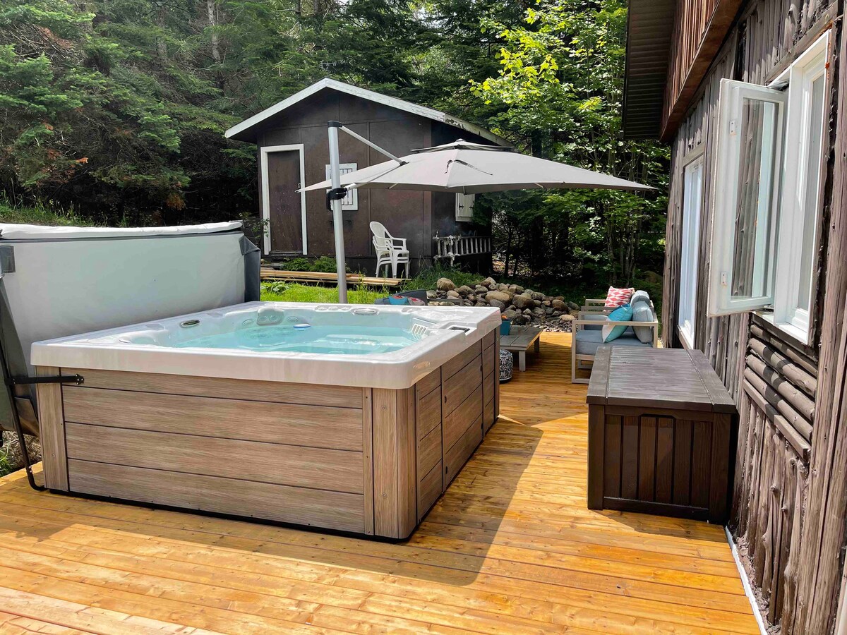 A wooden deck features a relaxing hot tub, surrounded by trees for added privacy. A shaded area is provided by a large umbrella, along with outdoor seating arrangements. The natural stone landscape adds a rustic charm to the space.