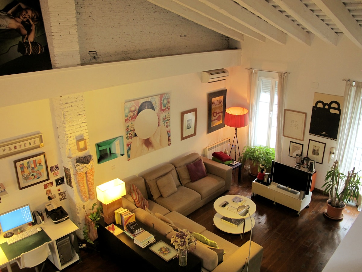 A spacious living area is highlighted by high ceilings with exposed beams and natural light from large windows. A comfortable sectional sofa sits alongside a coffee table, while artwork adorns the walls. Plants bring a touch of nature, contributing to the inviting ambiance.