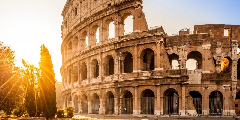 The Colosseum is showcased at sunrise, featuring its iconic arches and textured stone facade. Sunlight creates a warm glow, contrasting with the structure's weathered surface. Surrounding greenery adds a natural element to the historic scene, enhancing the monument's grandeur.