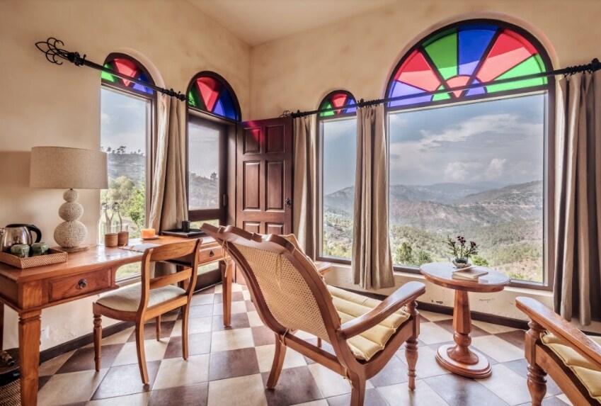 A cozy sitting area features large vintage windows with colored glass accents, allowing natural light to illuminate the interior. A wooden desk and two chairs complement the checkered floor, while a small circular table holds a decorative plant, all framed by scenic mountain views.