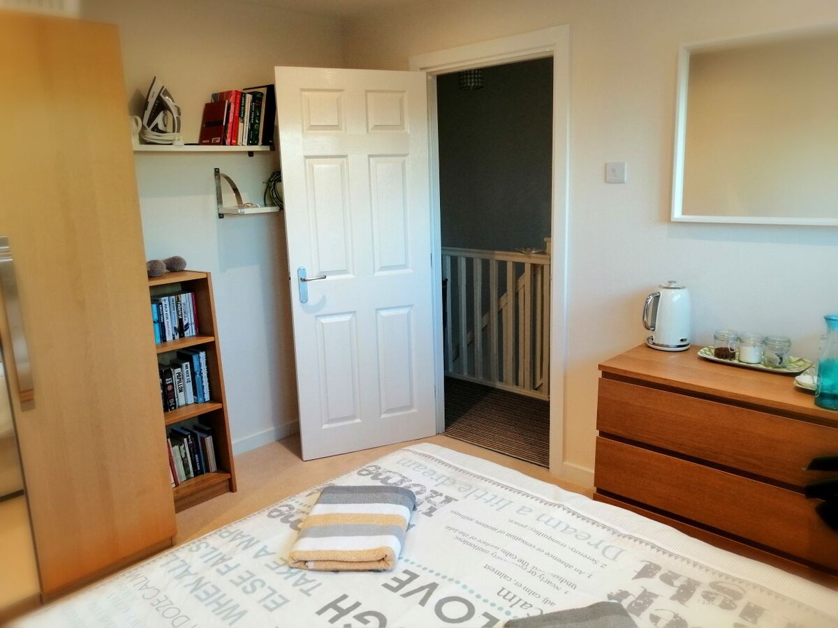 A clean and tidy double room features a spacious bed with patterned linens. A wardrobe holds books, while a kettle and snacks are arranged on a wooden dresser. A door leads to a stairway, enhancing accessibility to shared bathroom facilities.