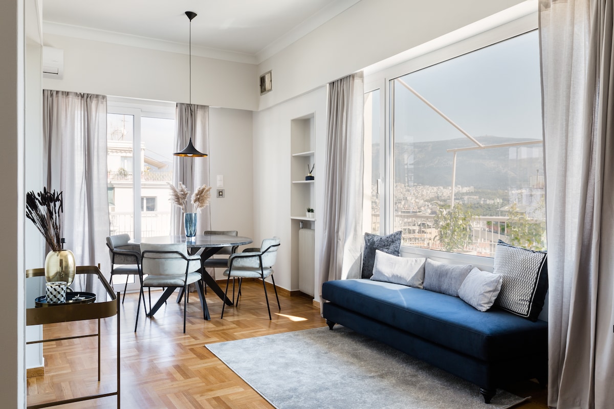 A luminous living area is showcased with a blue sofa and a wooden dining table surrounded by comfortable chairs. Large windows offer panoramic views of the city, while sheer curtains allow natural light to flood the space, enhancing its airy feel.