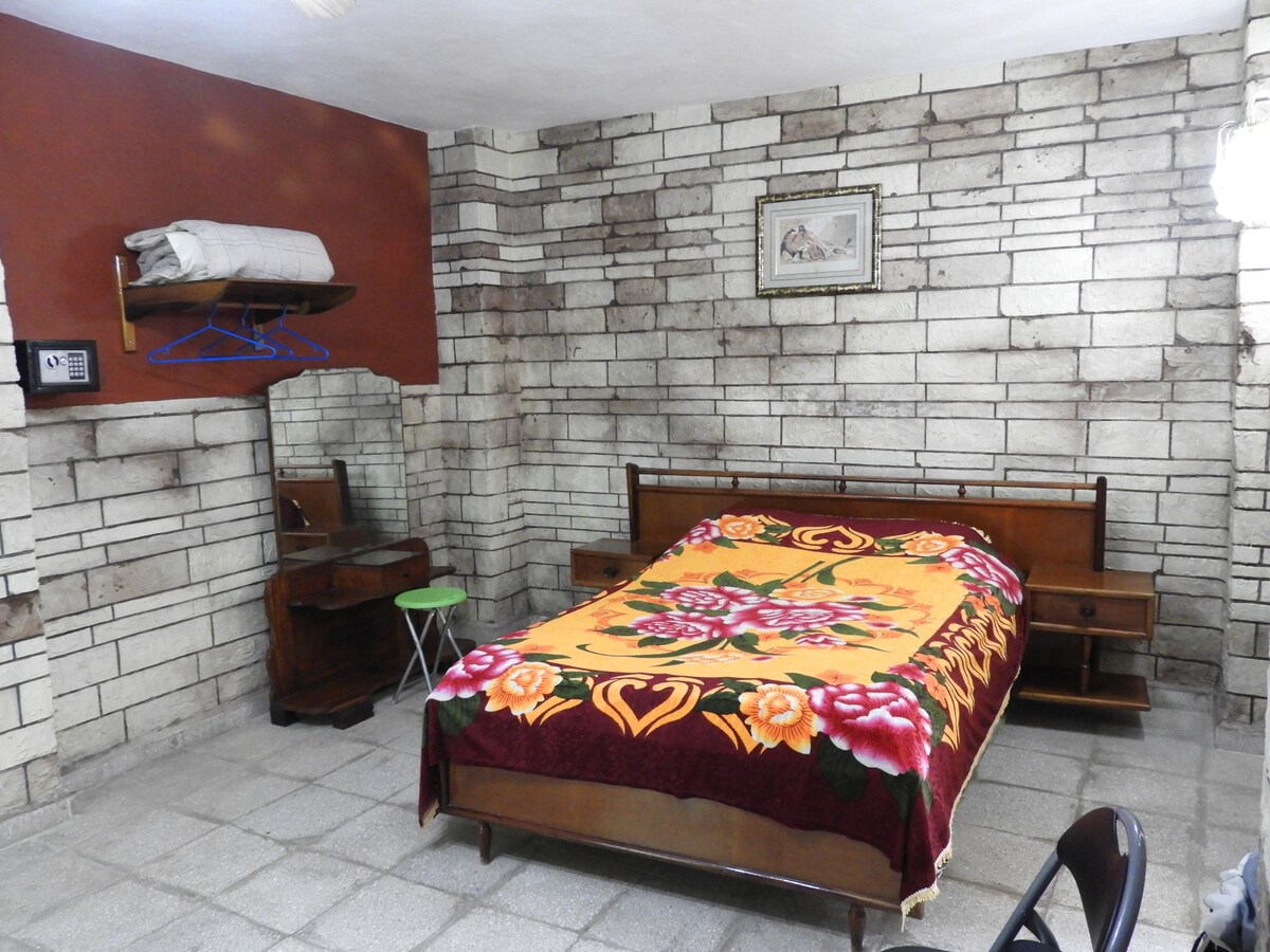 The room features textured stone walls and a spacious bed adorned with a floral blanket. A small green stool and a wooden nightstand complement the design. A wall shelf holds folded linens and a framed image adds a decorative touch.