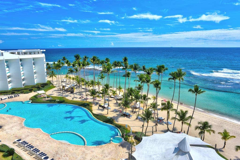 An aerial view captures the expansive pool area surrounded by palm trees and the sandy beach leading to the Caribbean Sea. The crystal-clear water of the pool contrasts with the vibrant blue of the ocean, creating a serene coastal atmosphere.