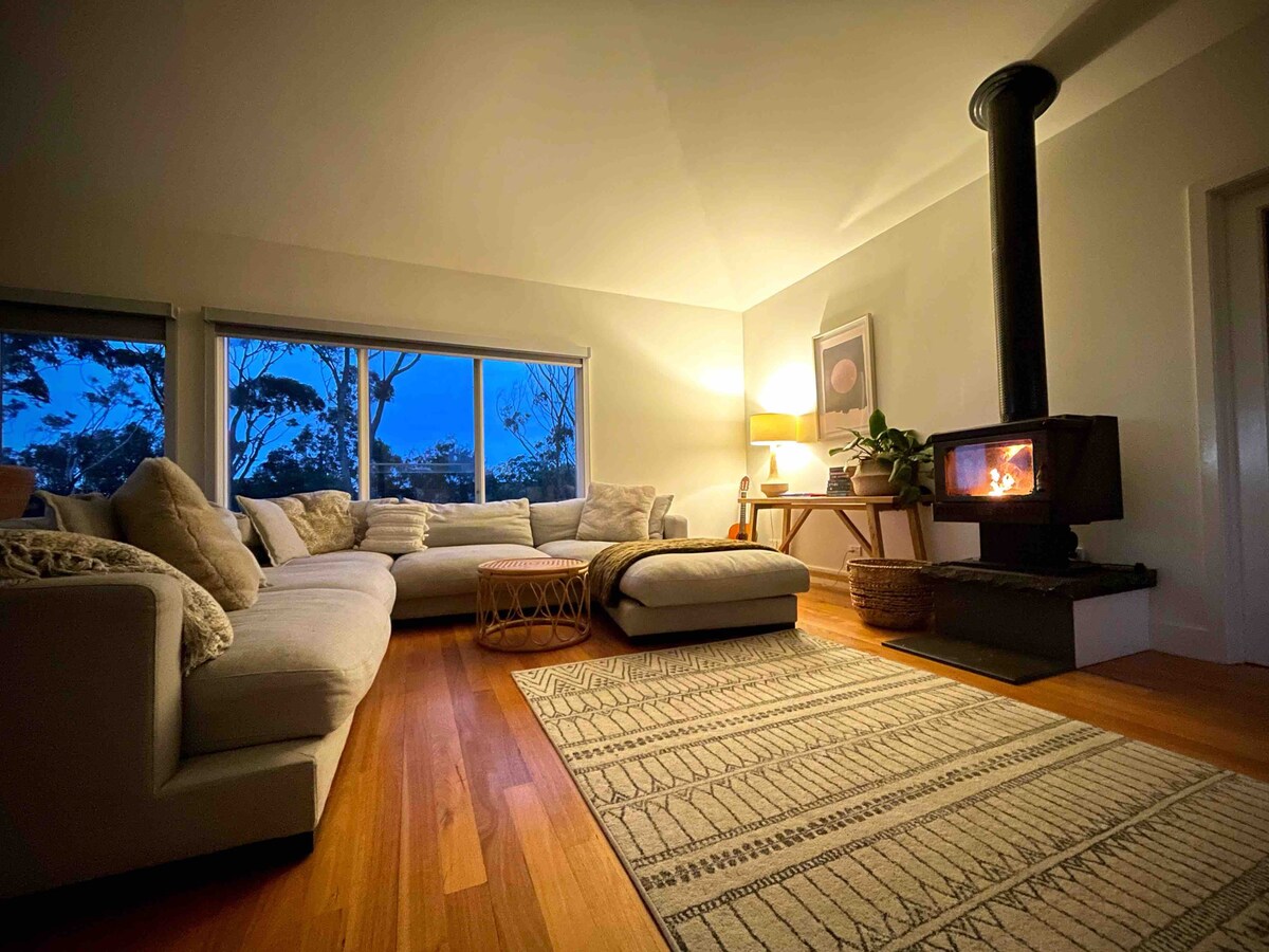 A spacious living area is illuminated by a warm glow from a wood heater, complementing the cozy aesthetic. A large sectional sofa surrounds a simple coffee table, while the large windows offer views of the outside. A softly patterned area rug enhances the inviting ambiance.