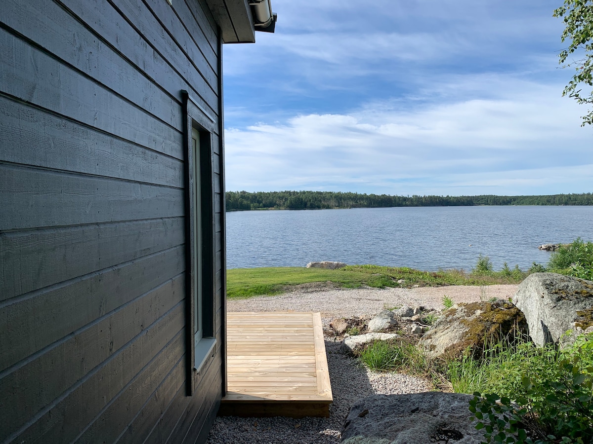 A side view of the Attefallshus is presented, showcasing the wooden deck leading to a serene lakefront. The calm waters are framed by lush greenery and distant trees, under a partly cloudy sky. Natural elements create a peaceful, outdoor setting.