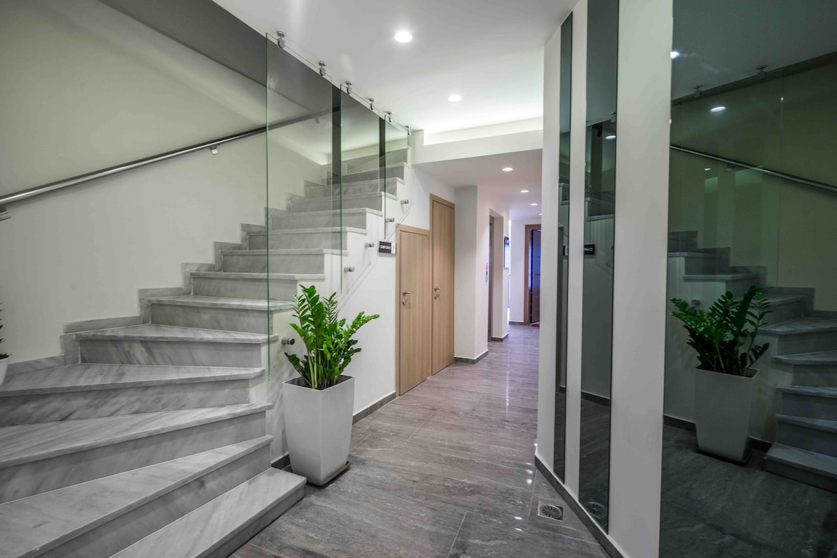 A sleek staircase is positioned at the center, flanked by glass panels that enhance the modern aesthetic. Potted plants add a touch of greenery, while the polished marble flooring reflects soft overhead lighting throughout the bright and airy hallway.
