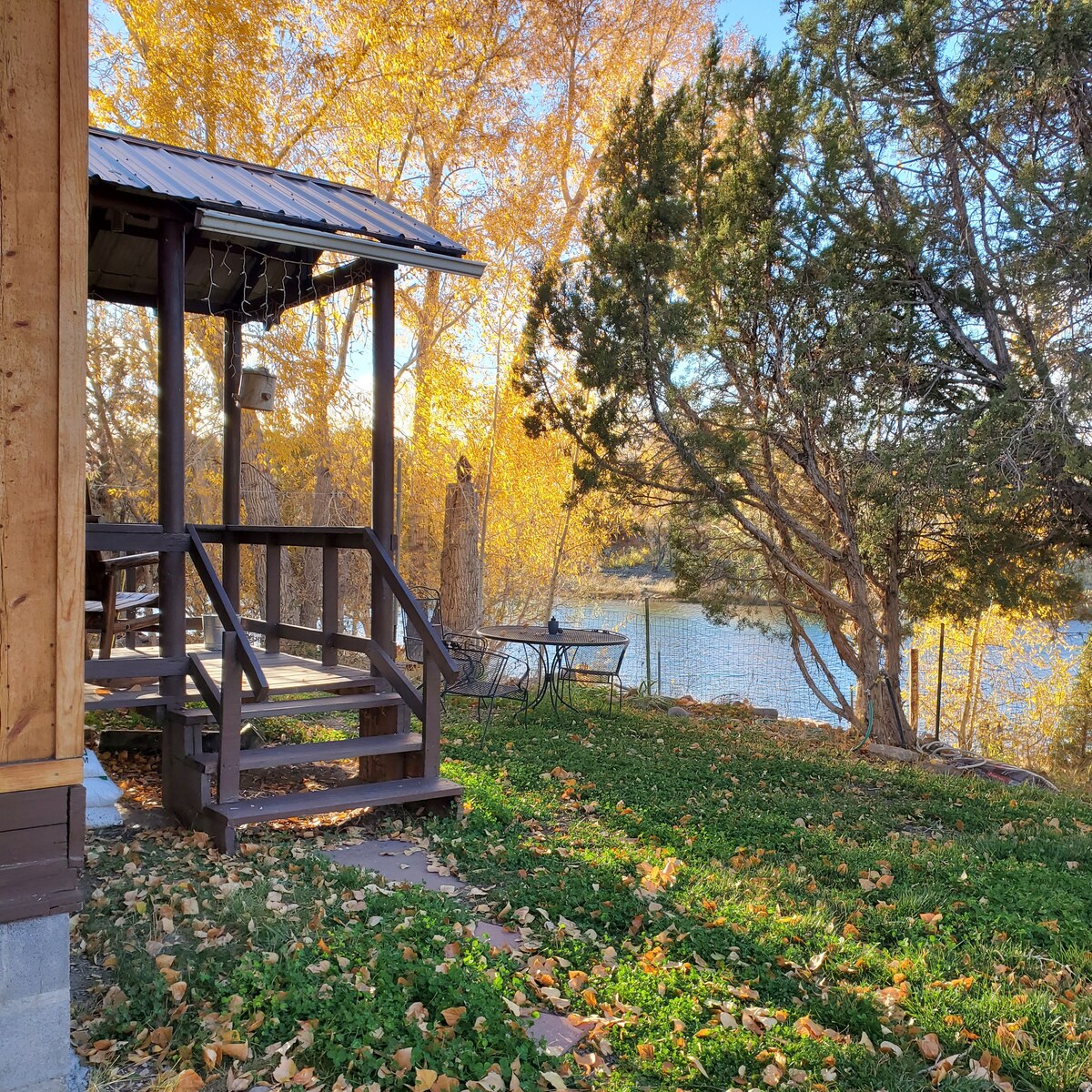 A porch leads to a shaded area with wooden steps, surrounded by vibrant autumn foliage. A picnic table is partially visible beside the water, with greenery and stones creating a natural border. Sunlight filters through the trees, illuminating the tranquil setting.