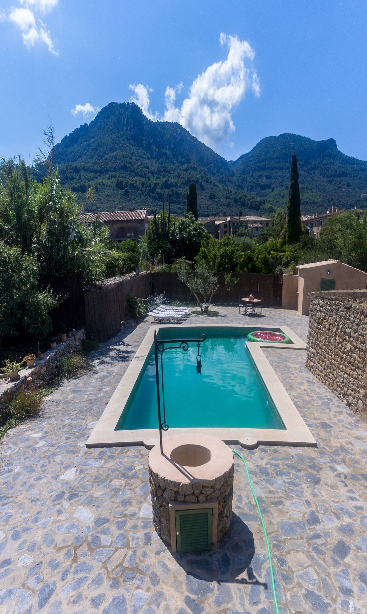 A tranquil pool area is visible, framed by lush greenery and natural stone walls. The swimming pool, surrounded by a stone terrace, reflects the clear blue sky and surrounding mountains. A shaded seating area is present nearby, offering a serene space for relaxation.