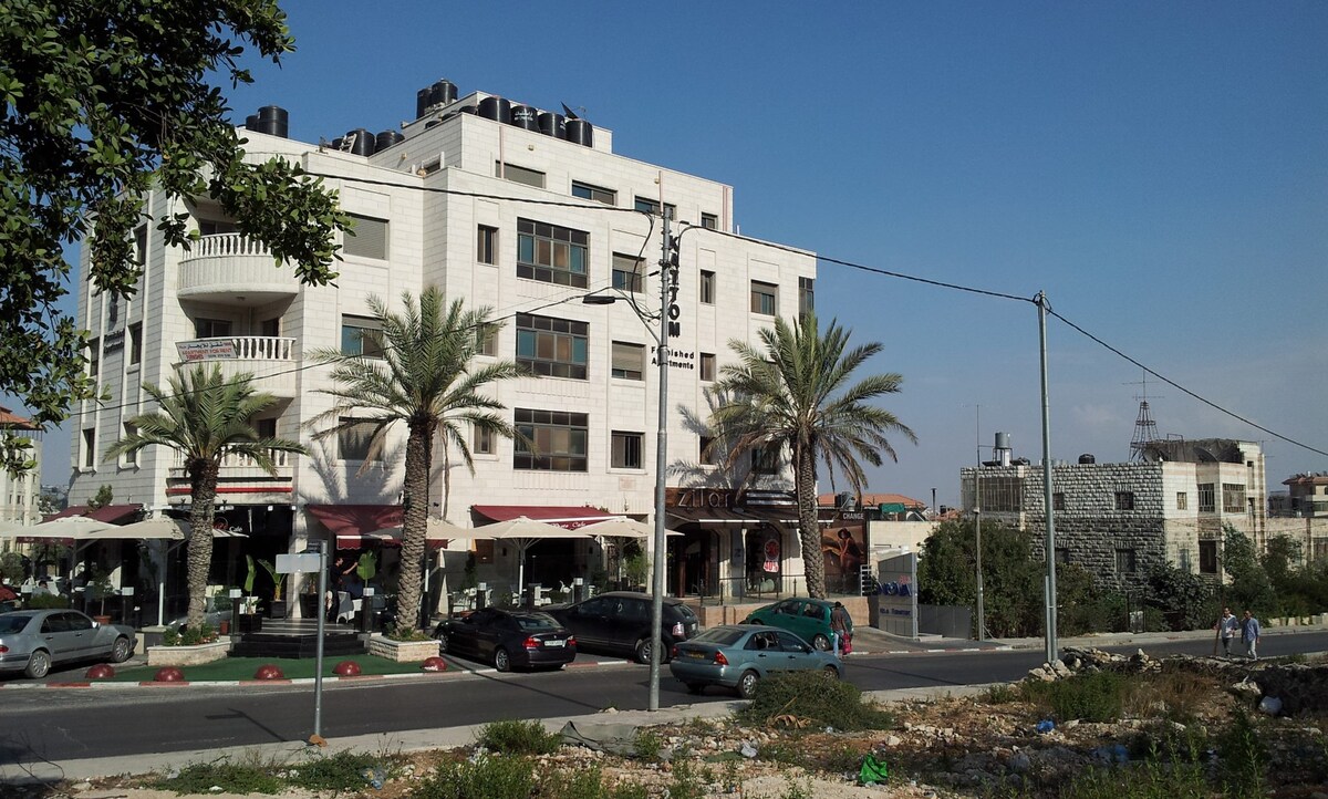 A several-story building is presented, featuring palm trees lining the sidewalk. The exterior includes a mix of stone and modern materials, with balconies visible on the upper floors. Restaurants with awnings are located at street level, and parked cars are seen along the road.