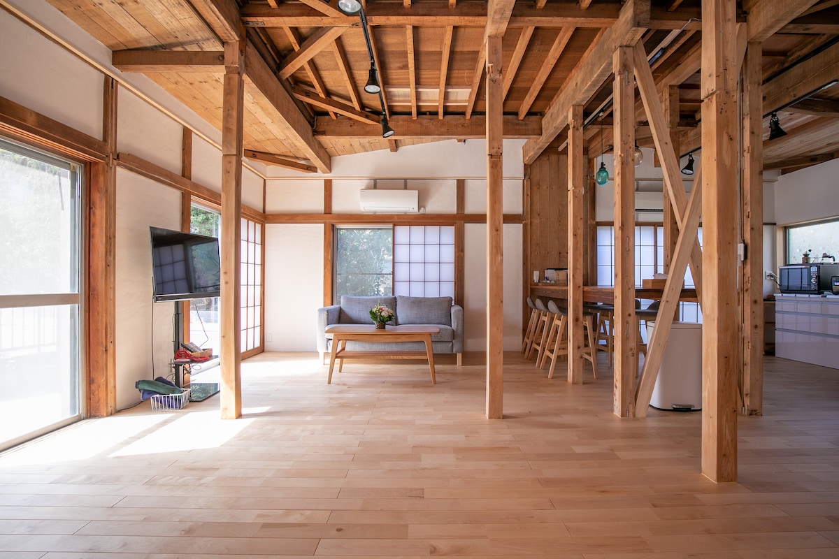An open living area features warm wooden beams and a light wooden floor. A comfortable sofa sits in front of a television, while a dining table is positioned nearby. Large windows provide ample natural light, illuminating the serene space.
