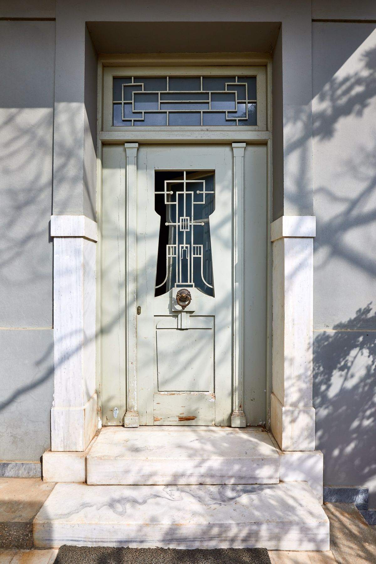 An elegant front door is framed by white marble pillars, showcasing intricate geometric designs in the glass. Shadows from nearby trees gently play across the muted exterior wall, enhancing the door's classic character.