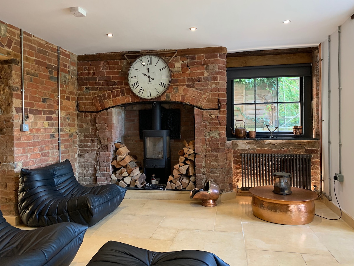 A rustic feature wall of exposed red brick hosts a large clock above a modern wood-burning stove. A stack of logs is neatly arranged beside it. Comfortable low-seating black sofas are placed in front of a round copper coffee table, with natural light filtering through the window.