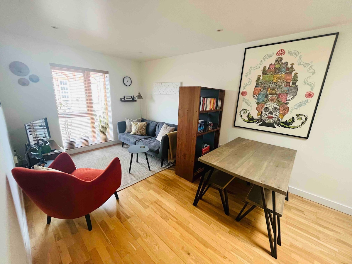 The living area features a red accent chair and a gray sofa, along with a wooden table and bookshelves filled with books. Light enters through a large window, illuminating the wooden floor and artwork on the wall.