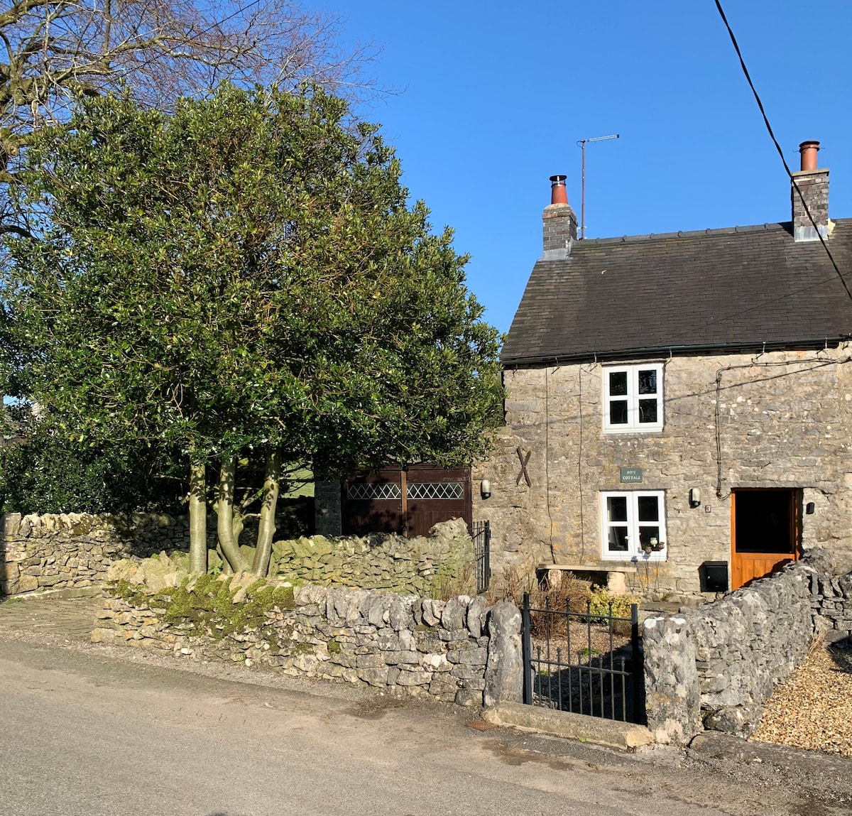 The exterior of Rose Cottage is presented, showcasing its traditional stone façade and steep roof. A large tree is visible in the front garden, surrounded by a stone wall and gated entrance, with a clear blue sky above.