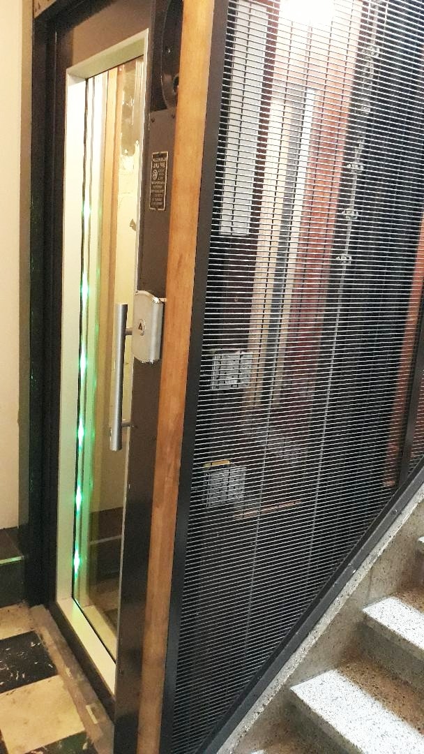 An antique elevator is shown with a wooden frame and glass doors, featuring illuminated lights along the edges. The surrounding area displays decorative tile flooring and a staircase leading to the upper levels.