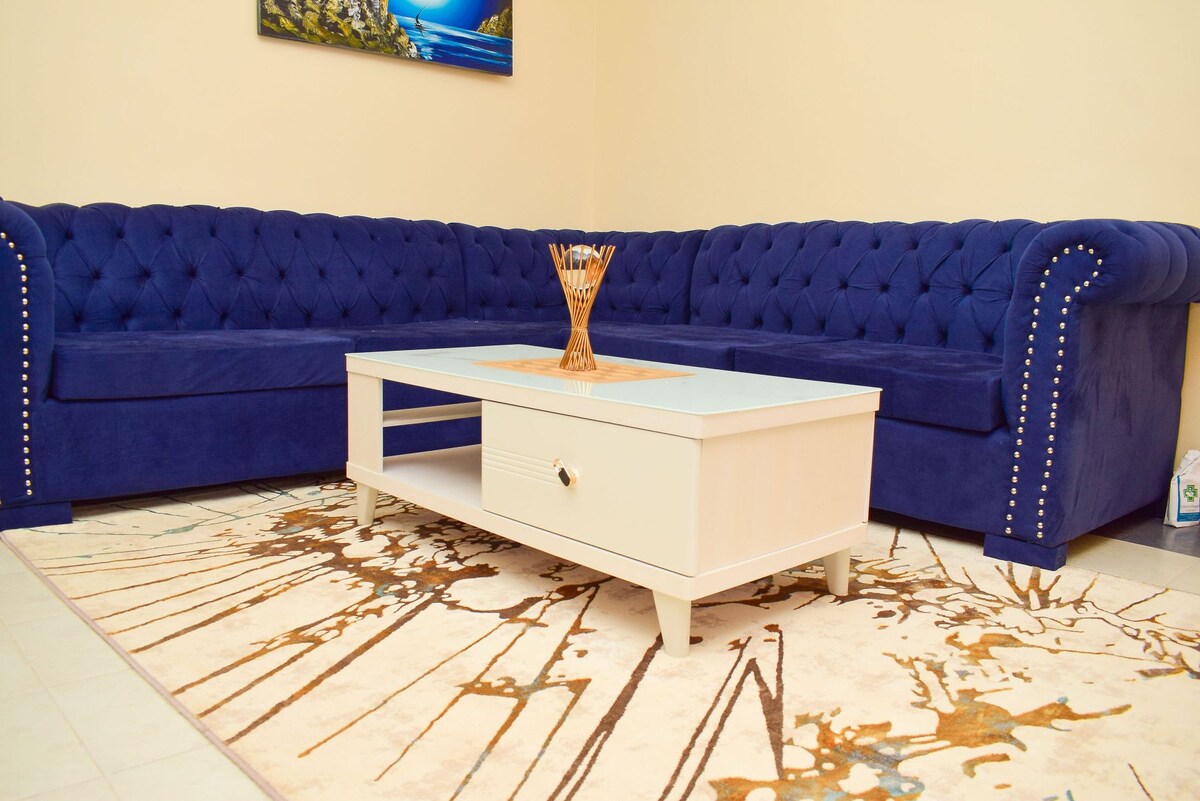 A plush blue sectional sofa with tufted detailing provides ample seating. A central white coffee table with an open shelf is positioned on a patterned area rug, featuring abstract designs in earthy tones. A piece of wall art depicting a serene landscape is displayed above.