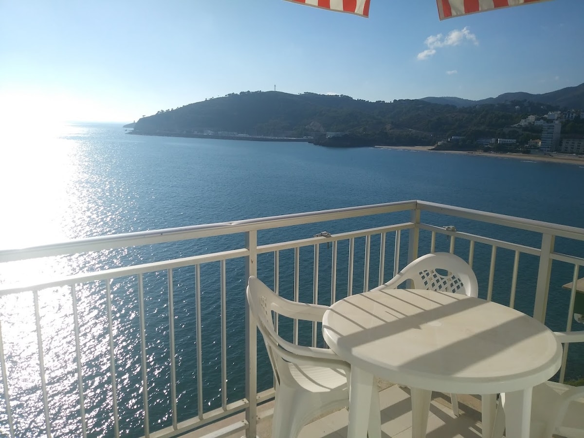A balcony offers sweeping views of the shimmering ocean, with gentle waves visible in the distance. A small round table is accompanied by two white chairs, creating a peaceful outdoor space for relaxation. Bright sunlight reflects off the water, enhancing the coastal atmosphere.