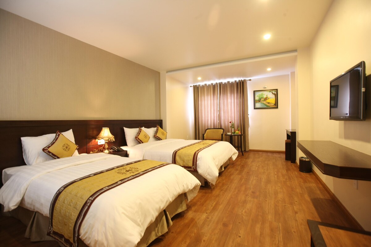 A spacious room features two beds with neutral bedding and decorative pillows. A wooden desk and chair are positioned near large windows dressed with light curtains. A wall-mounted television is visible, along with framed artwork adding subtle color to the setting.