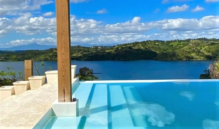 The private infinity pool extends towards a panoramic view of the lake and surrounding hills. Wooden beams frame the scene, with planters positioned along the edge. The sky is dotted with fluffy clouds, reflecting the calm waters below.
