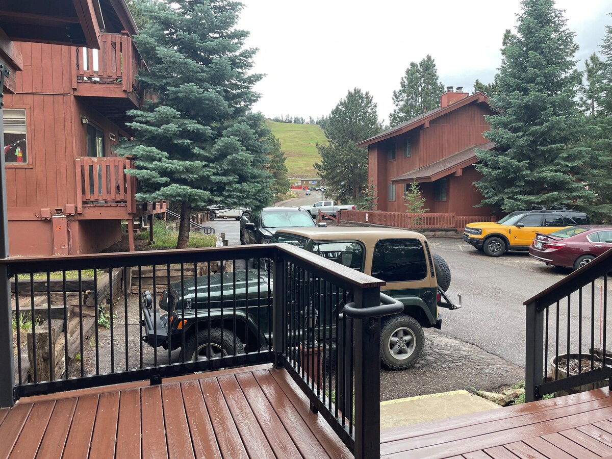 The view from the deck captures a quiet outdoor space, featuring a wooden railing and a landscaped area with evergreen trees. Several vehicles are parked in the lot, providing accessibility to the nearby buildings nestled among the trees.