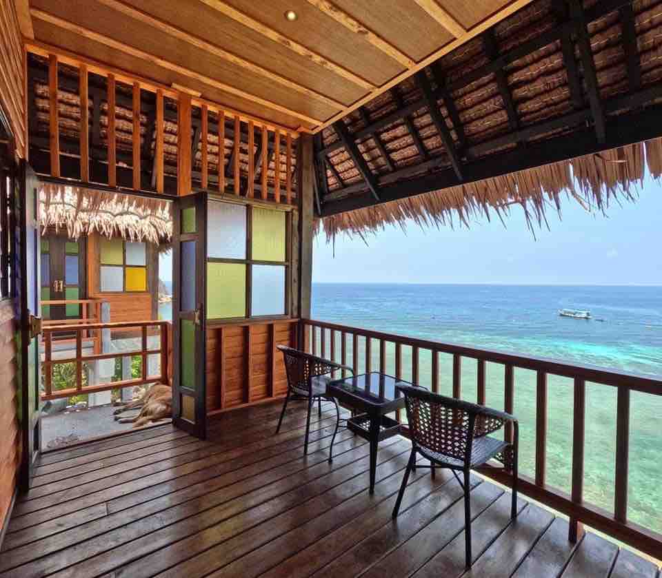 A spacious private balcony offers stunning views of the ocean, featuring wooden flooring and a thatched roof. Two wicker chairs and a small table provide a relaxing spot to enjoy the scenery. The entrance to the villa is framed by colorful glass panels.