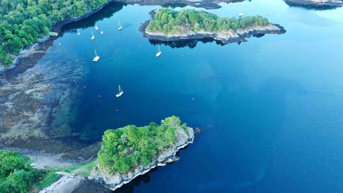 Aerial view over a tranquil body of water, showcasing small islands covered in greenery and scattered sailboats. The shoreline displays a mix of rocky and sandy areas, with the serene blue of the water reflecting the surrounding landscape.