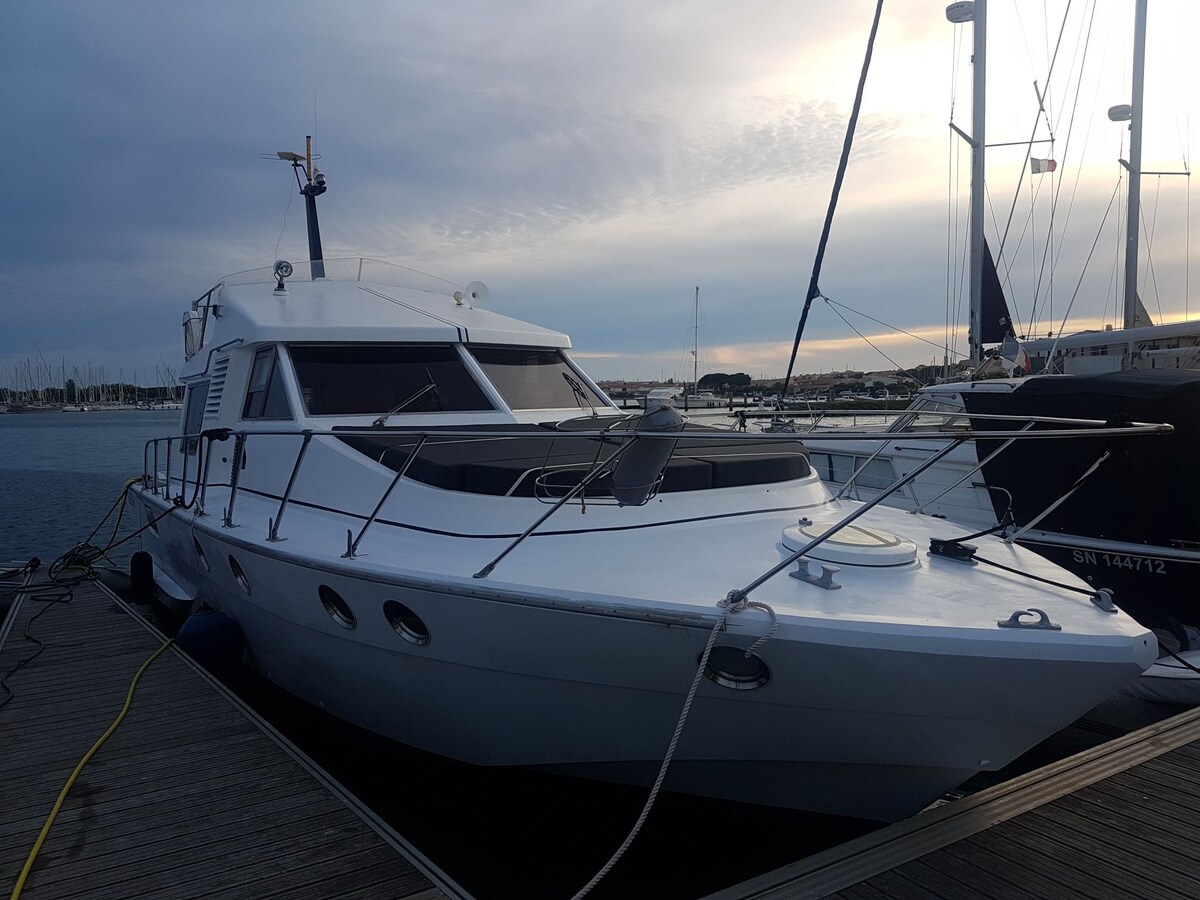 The exterior of the boat is showcased, featuring a sleek hull and spacious deck area. The design is modern, with prominent windows allowing natural light. Surrounding the boat are other vessels and a calm harbor, indicating a peaceful waterfront location.