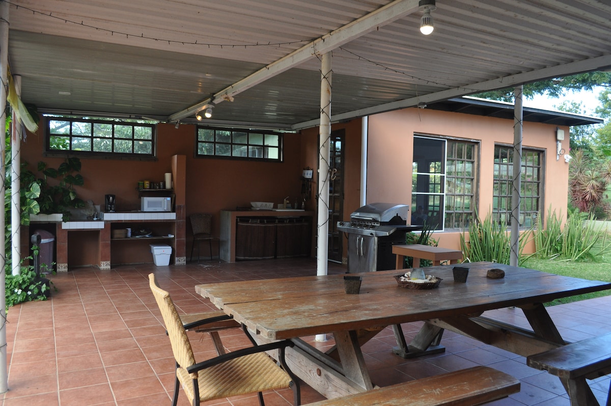 An outdoor dining area features a large wooden table surrounded by chairs. A grill and outdoor kitchen space are visible, with shelving, a microwave, and sink nearby. Natural greenery frames the area, and large windows provide views of the surrounding yard.