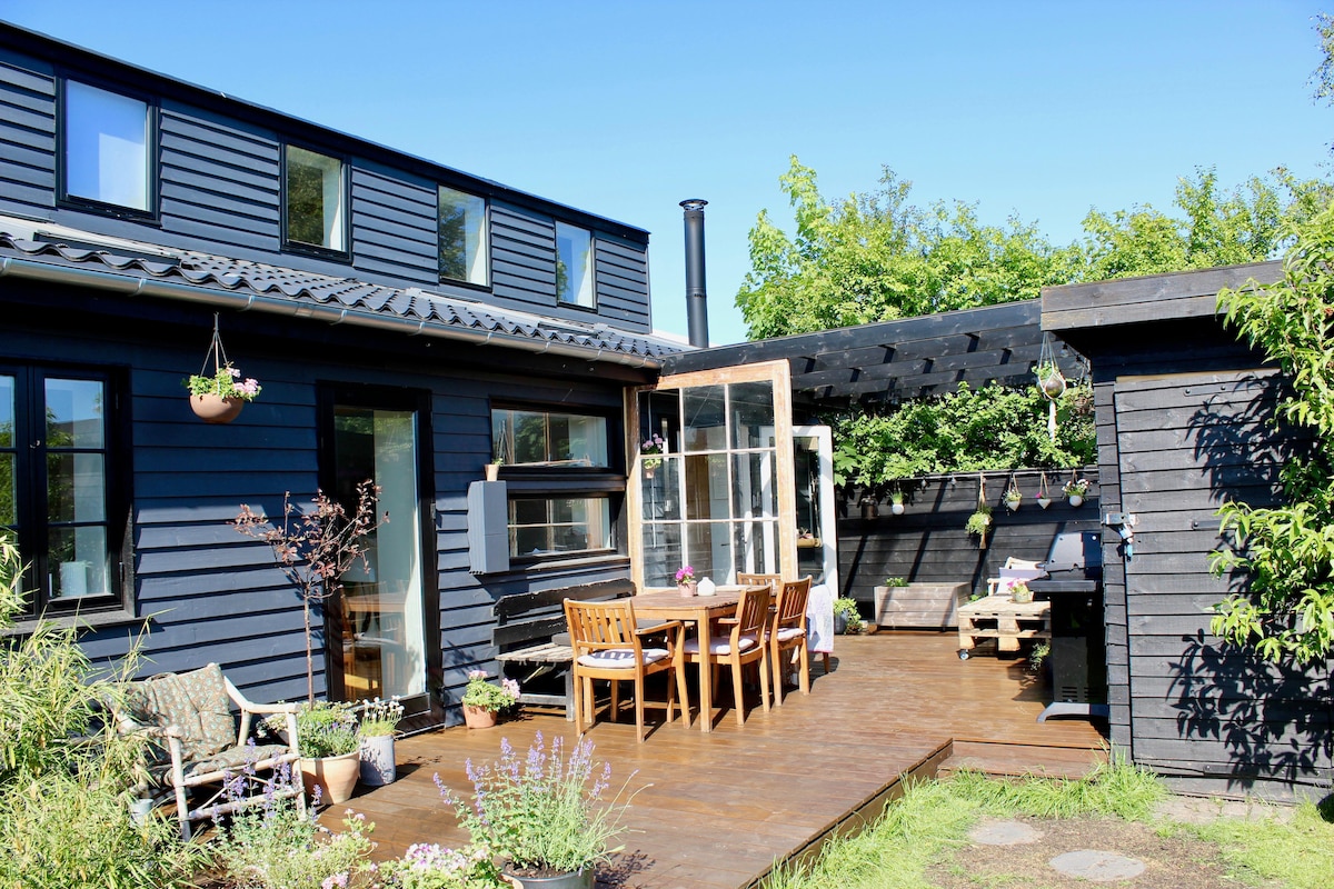 A cozy outdoor deck features a wooden dining table surrounded by several chairs, with vibrant greenery in pots enhancing the space. Large glass doors open to the interior, while a charming outbuilding is visible, showcasing a relaxed and inviting outdoor atmosphere.