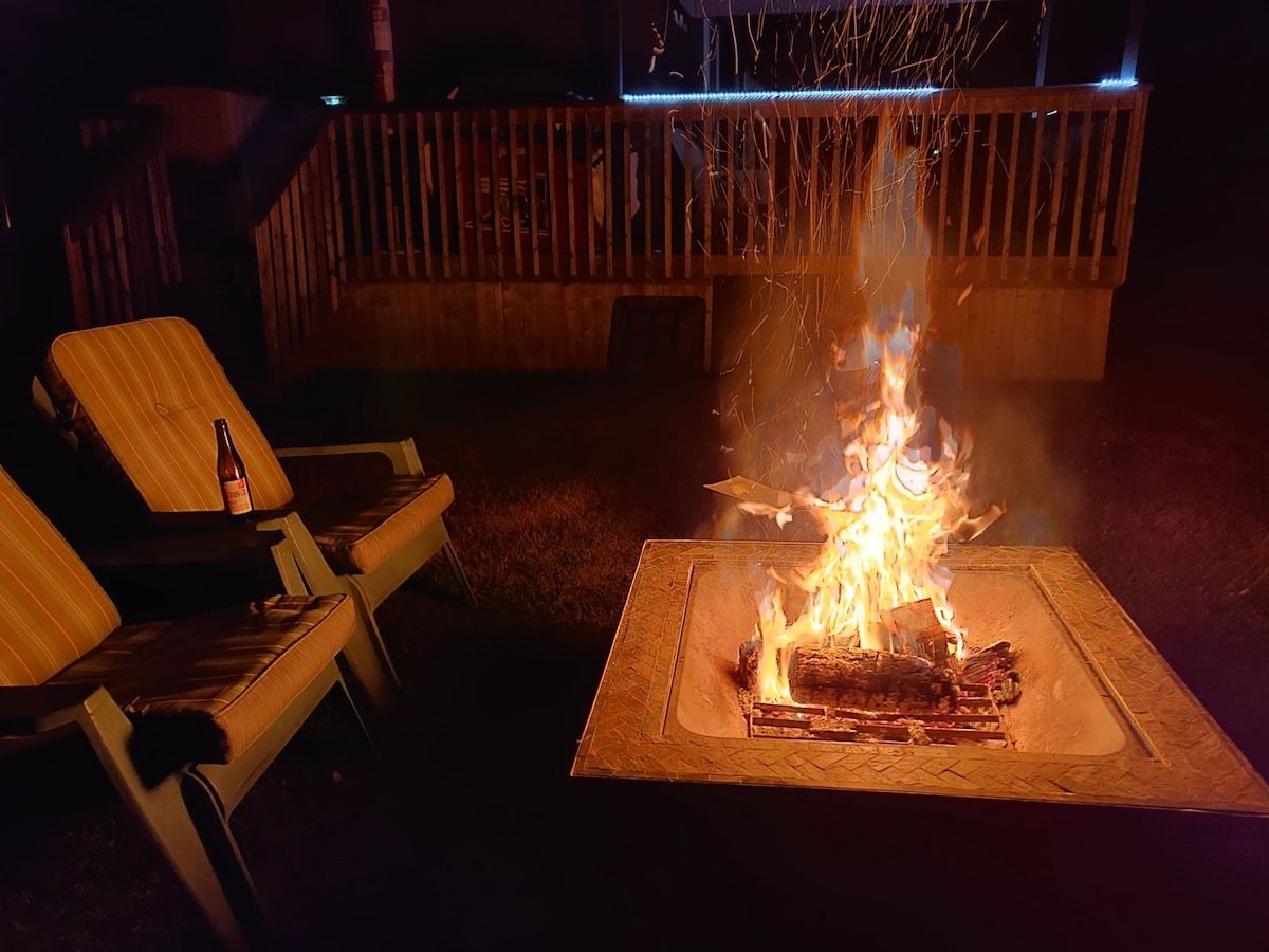 A fire pit is centrally positioned, with flames dancing above a bed of logs. Two striped outdoor chairs are placed nearby, providing a cozy setting for gathering. The background features a wooden deck illuminated softly, enhancing the evening ambiance.